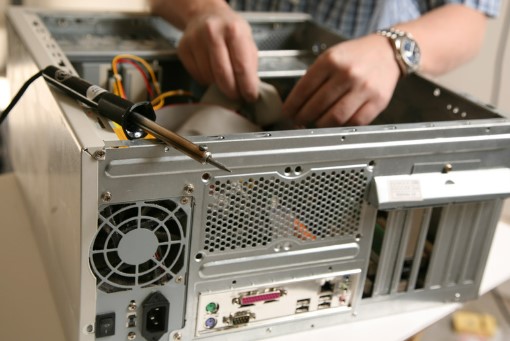 Person assembling or repairing a desktop computer, with hands inside the open case and various cables visible; a soldering iron is placed nearby.