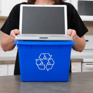 A person places a laptop into a blue recycling bin in an office setting.