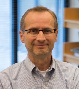 A man with glasses and short light hair, wearing a light blue collared shirt, is smiling in an office with shelves and windows in the background.