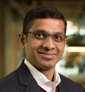 A man wearing glasses and a suit jacket smiles at the camera in an indoor setting with blurred lights in the background.