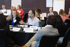A group of people sit around conference tables with state name cards, as one man speaks into a microphone during a meeting.
