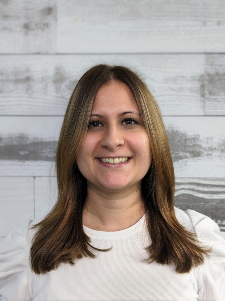 A person with long brown hair smiles in front of a wood-paneled background.