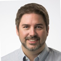 A man with short brown hair, a trimmed beard, and a light plaid shirt smiles at the camera against a plain white background.
