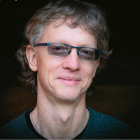 A middle-aged man with wavy light brown hair, glasses, and a slight smile, wearing a dark green shirt, poses in front of a dark background.