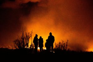 Silhouettes of four people stand in front of a large, intense fire, with smoke and flames illuminating the night sky.