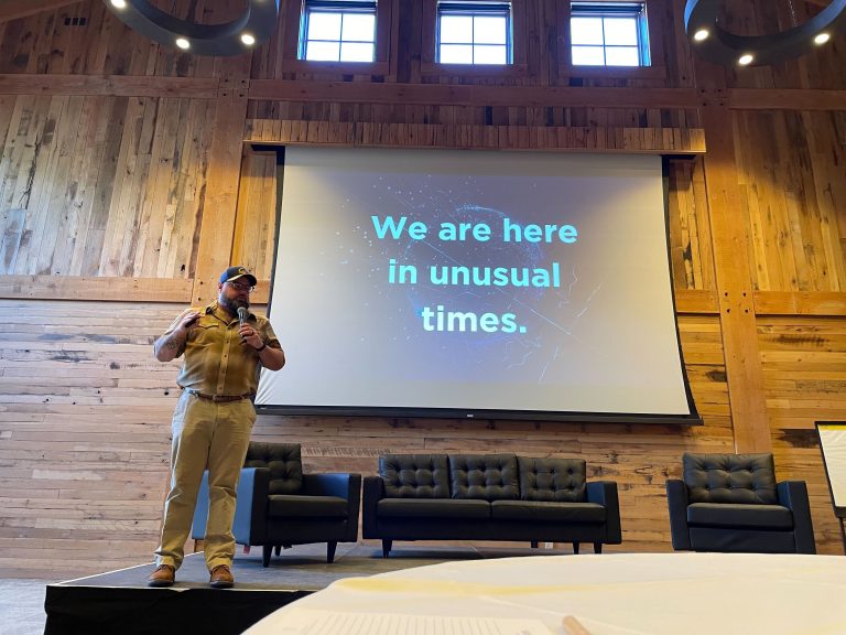 A person in casual attire stands on a stage in front of a screen displaying the words "We are here in unusual times" inside a wooden-panelled room.