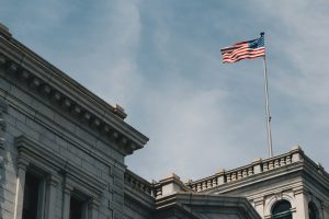 An American flag flies atop a flagpole on the roof of a classical stone government building under a partly cloudy sky.