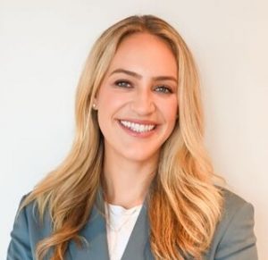 A woman with long blonde hair wearing a light gray blazer and white top smiles at the camera against a plain light background.