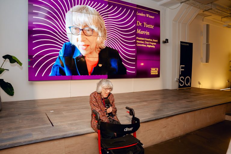 An older woman speaks into a microphone while sitting with a walker in front of a screen displaying her photo and an award announcement.