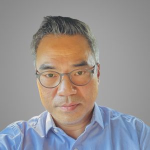 Headshot of a middle-aged man with short gray hair and glasses sits in a car, wearing a blue collared shirt. Sunlight reflects off his glasses.