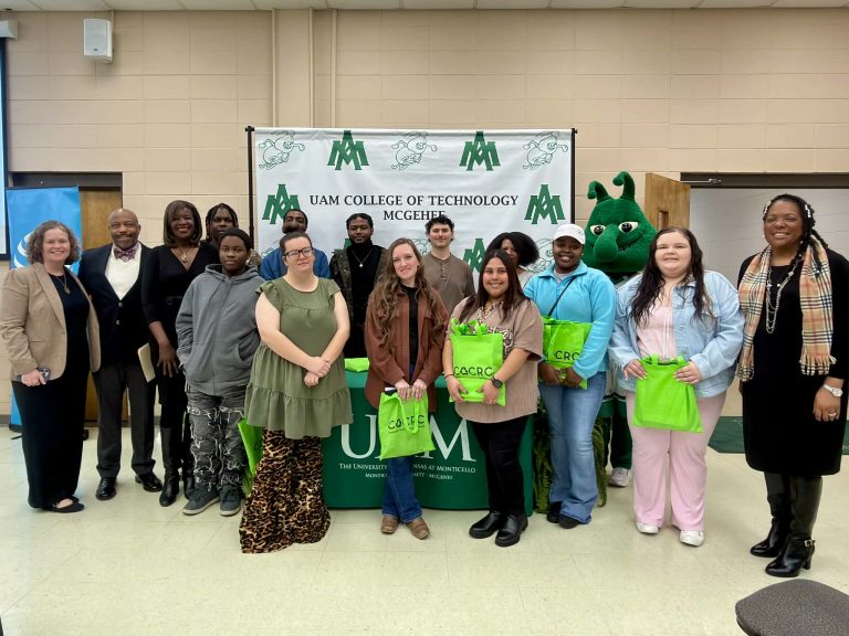 Group of students and staff posing indoors in front of a UAM College of Technology–McGehee banner and table display, several holding bright green tote bags, with a green school mascot standing behind them.