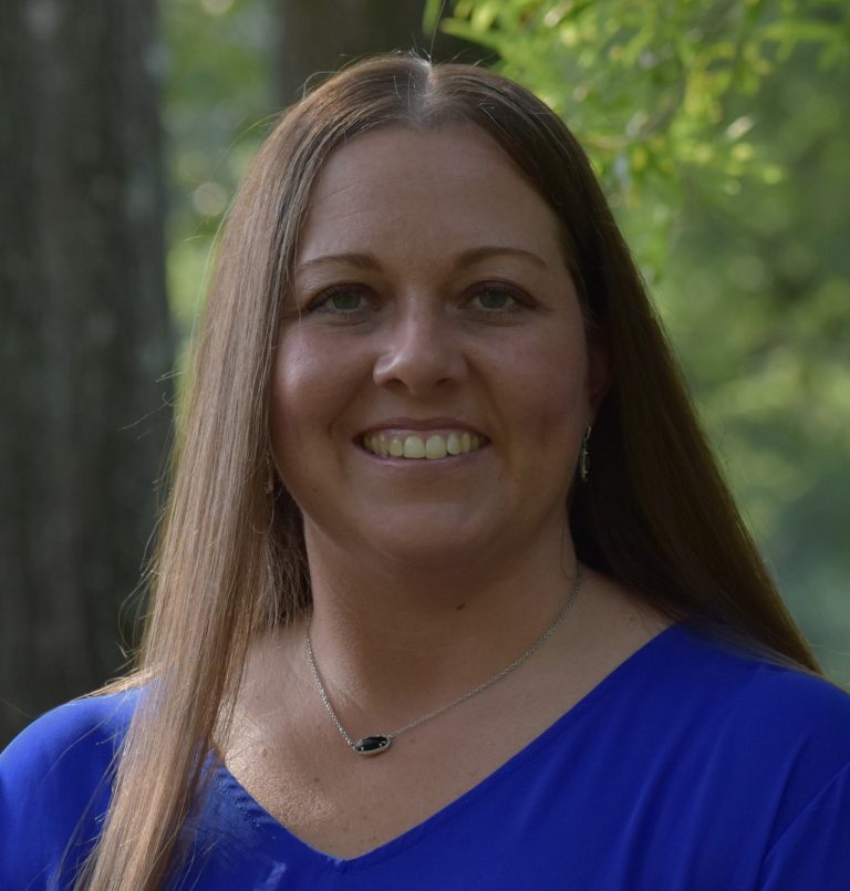 Head-and-shoulders portrait of a smiling woman with long brown hair wearing a blue top and a necklace, photographed outdoors with trees and soft greenery in the background.