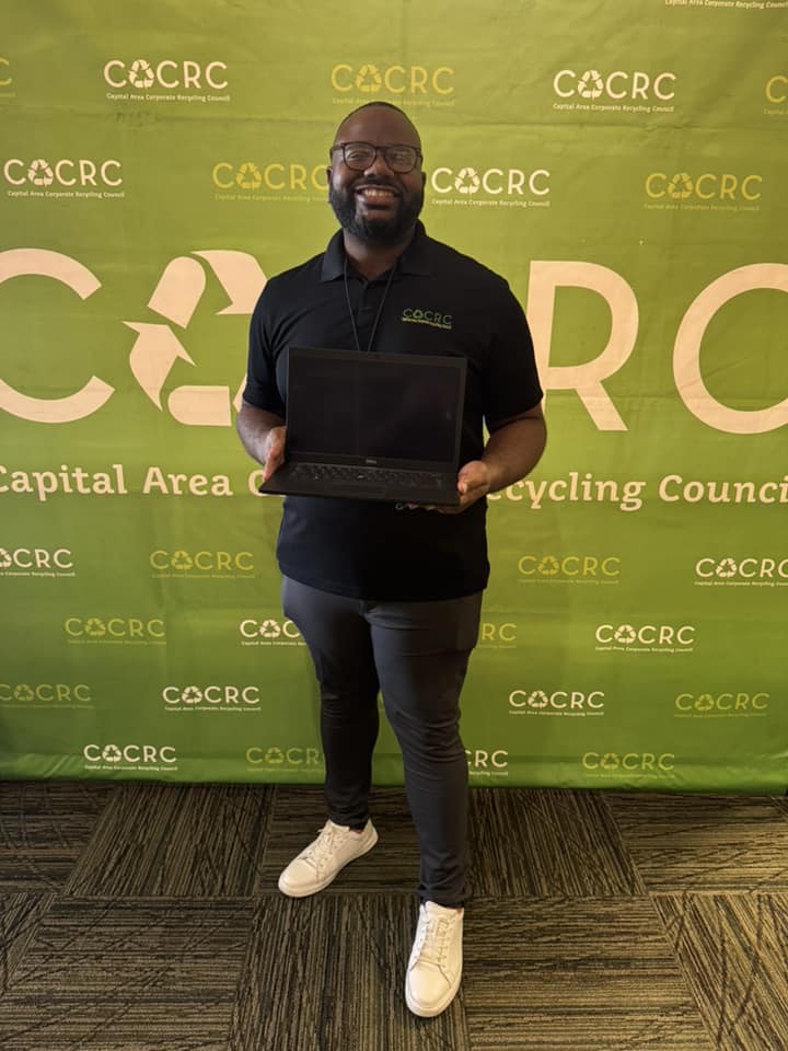 A man stands in front of a green CACRC backdrop, holding an open laptop and smiling, dressed in a black polo and sneakers.