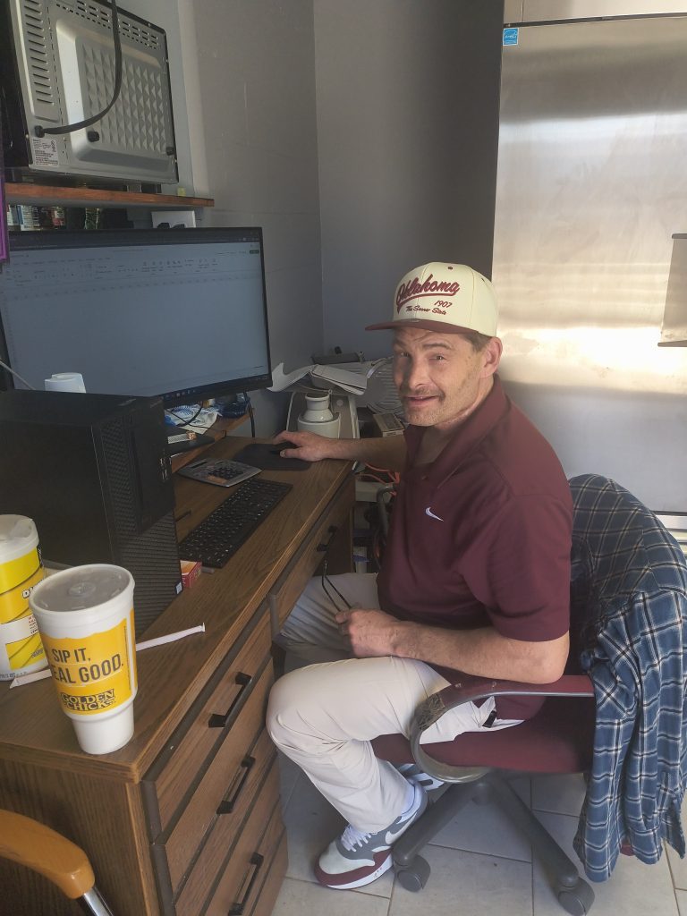 A man wearing a maroon polo shirt and an "Oklahoma" baseball cap sits at a wooden desk using a computer with a spreadsheet open on it.