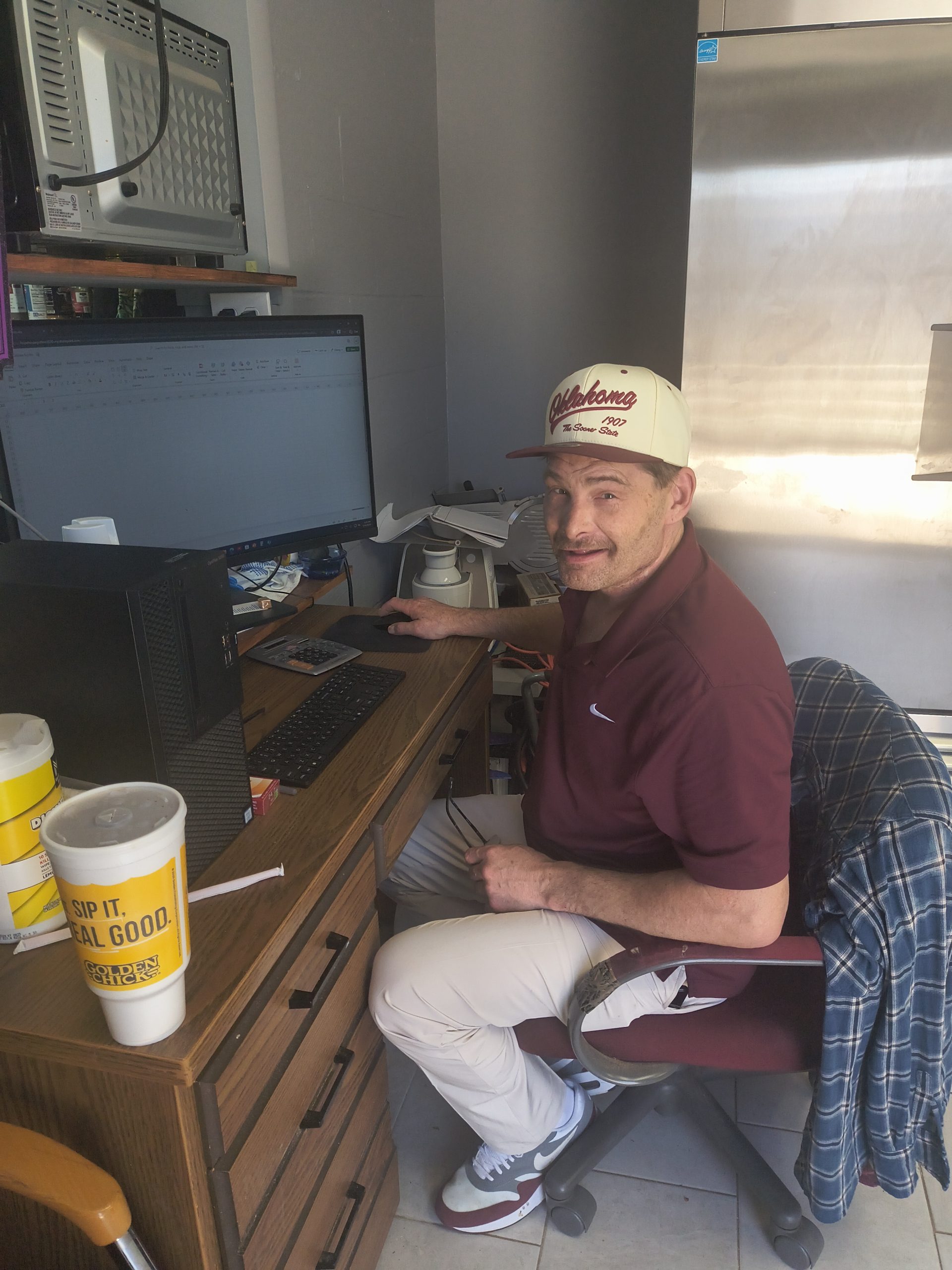 A man wearing a maroon polo shirt and an "Oklahoma" baseball cap sits at a wooden desk using a computer with a spreadsheet open on it.