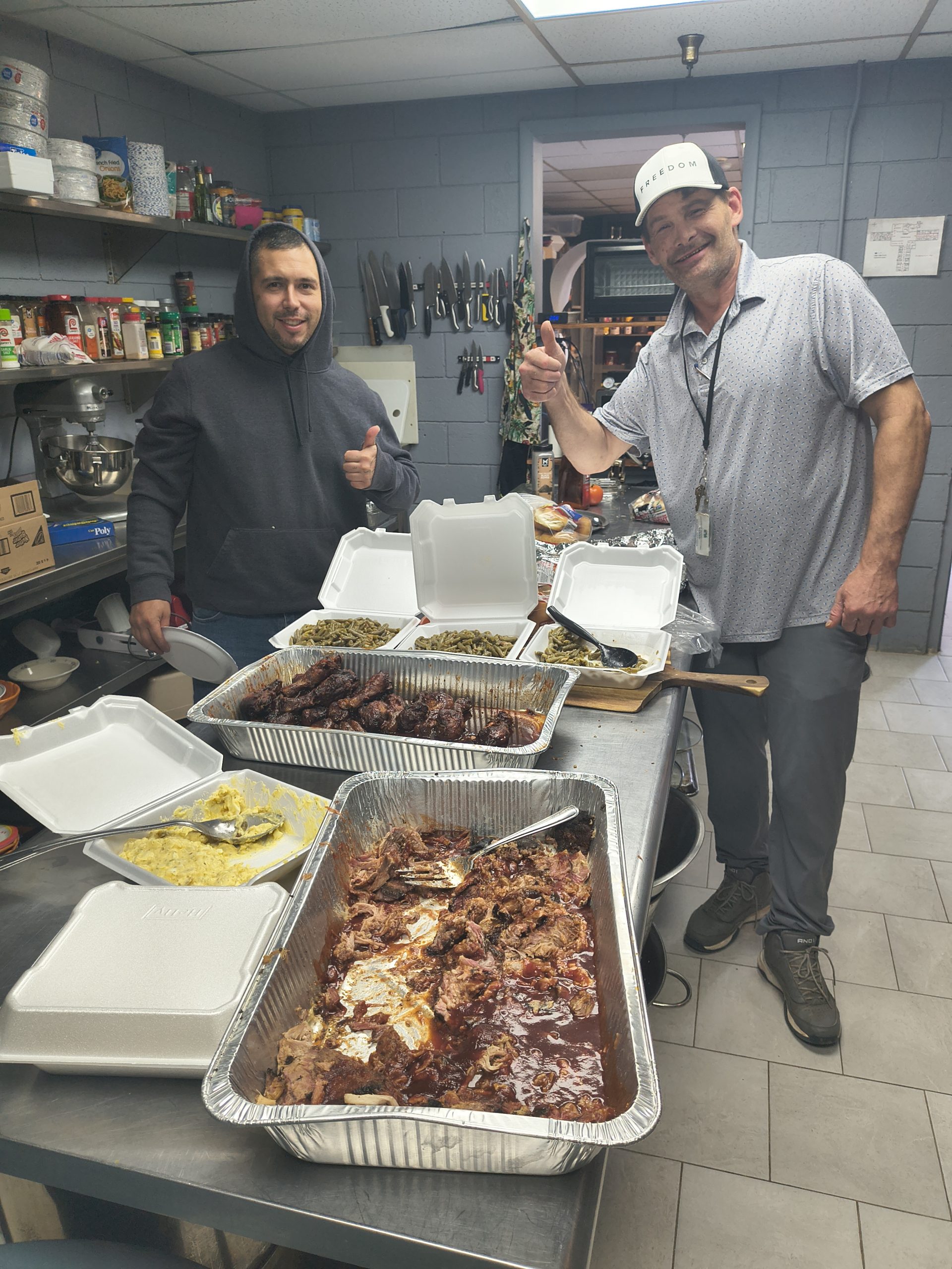 Two men standing in a commercial kitchen giving thumbs-up signs behind a counter filled with large catering trays of food, including pulled pork, BBQ chicken, green beans, and corn.