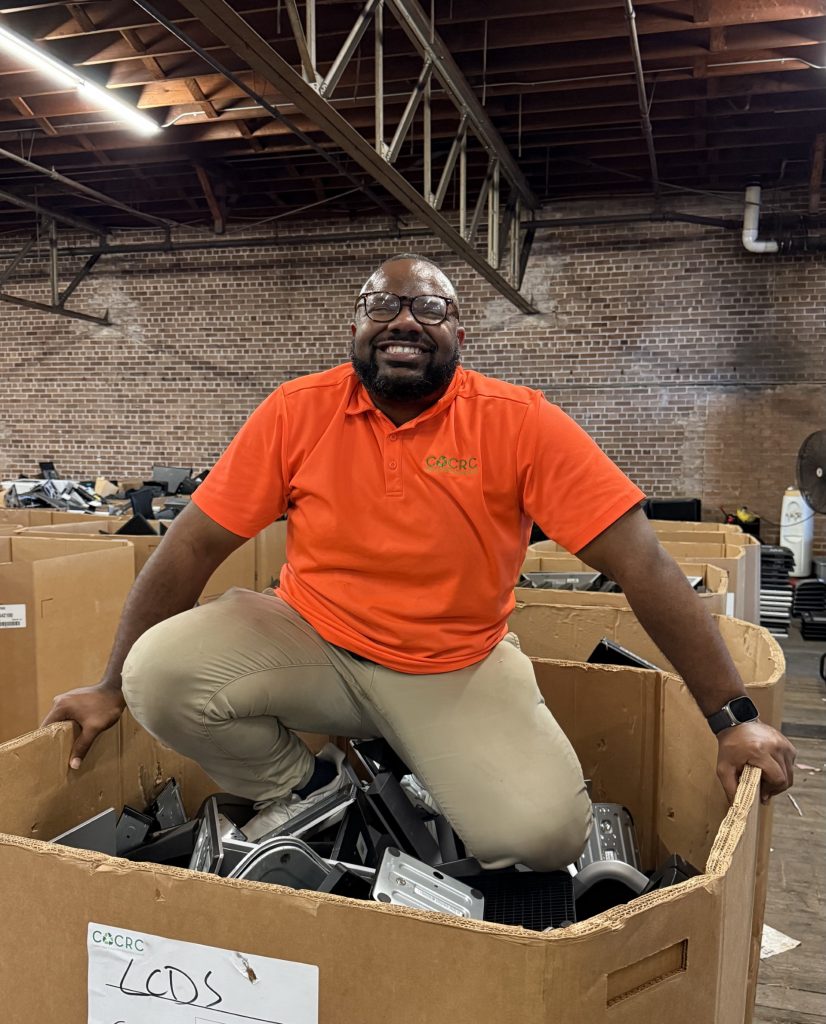 Man wearing an orange CACRC polo kneels on top of a large bin filled with recycled computer parts inside a warehouse, smiling at the camera.