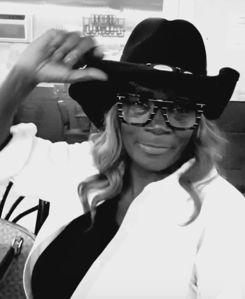 Black-and-white portrait of a woman wearing a cowboy hat, patterned glasses, and a light-colored jacket while smiling and adjusting the brim of her hat indoors.