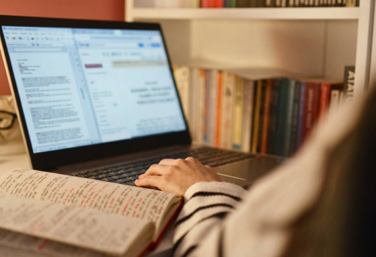 Person using a laptop beside an open notebook filled with handwritten notes in a home study space