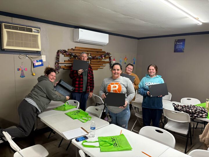 Five adults in a community classroom smile while holding open laptops, with tables, chairs, and bright green tote bags in the foreground.