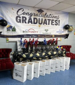 Graduation celebration setup with a “Congratulations Graduates!” banner featuring Neighborhood Hope Dealers, Digitunity, Human-I-T, and AT&T Connected Learning logos, with gift bags and graduation-themed decorations displayed below.