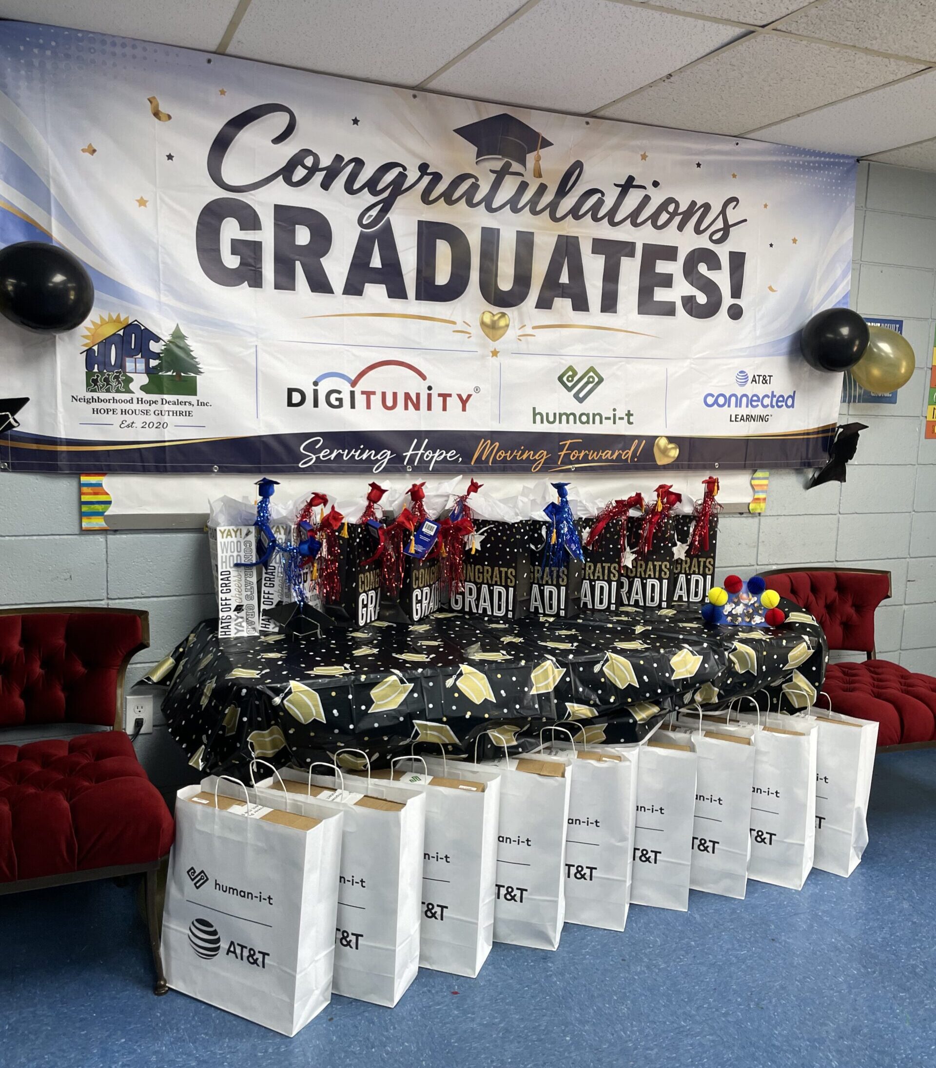 Graduation celebration setup with a “Congratulations Graduates!” banner featuring Neighborhood Hope Dealers, Digitunity, Human-I-T, and AT&T Connected Learning logos, with gift bags and graduation-themed decorations displayed below.