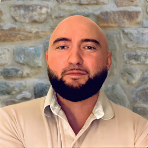 Headshot of a man with a shaved head and dark beard wearing a light collared shirt against a stone wall background.