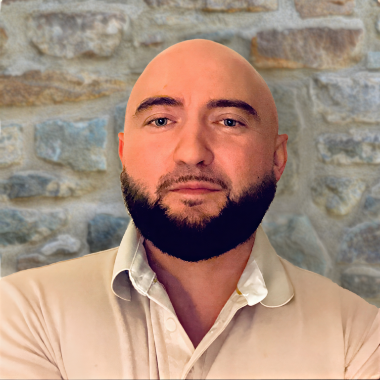 Headshot of a man with a shaved head and dark beard wearing a light collared shirt against a stone wall background.