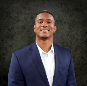 Kamelle Gomez smiling in a navy suit jacket and white shirt against a dark textured background.