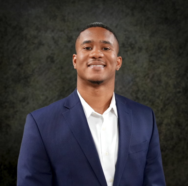 Kamelle Gomez smiling in a navy suit jacket and white shirt against a dark textured background.