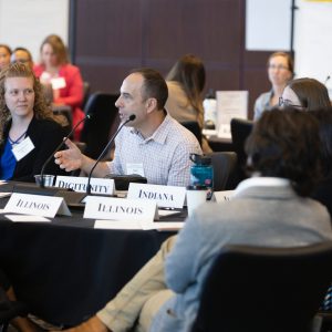 A group of people sit around conference tables with state name cards, as one man speaks into a microphone during a meeting.