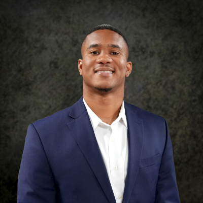 Kamelle Gomez smiling in a navy suit jacket and white shirt against a dark textured background.