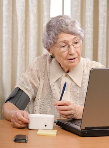 An elderly woman with a blood pressure cuff checks her laptop, holding a pen and a cup, seated at a table with a phone and sticky notes.